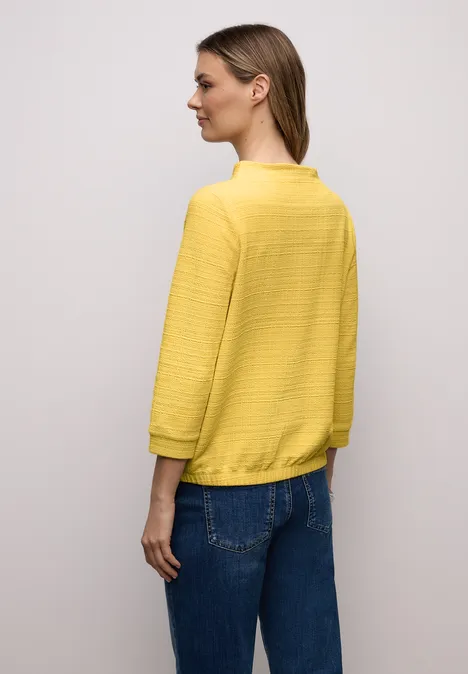 Eine Frau trägt einen gelben Pullover und blaue Jeans, von hinten vor hellem Hintergrund.